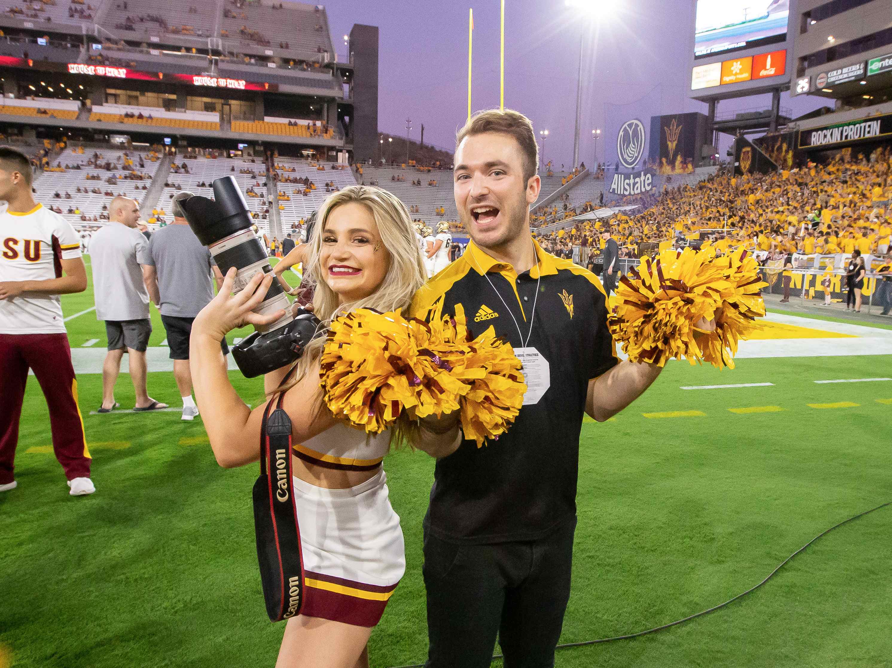 Billy Hardiman holding pom-poms with an ASU cheerleader holding his camera on the sidelines at Mountain America Stadium